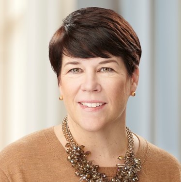 A person with short brown hair and a warm smile wears a brown top and an ornate necklace, set against a softly lit, neutral background.