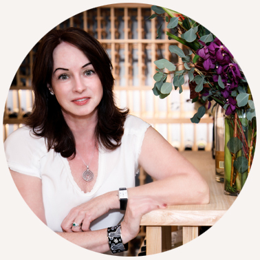 A person with dark hair, wearing a white top and a necklace, sits at a wooden table with a vase of purple flowers nearby.