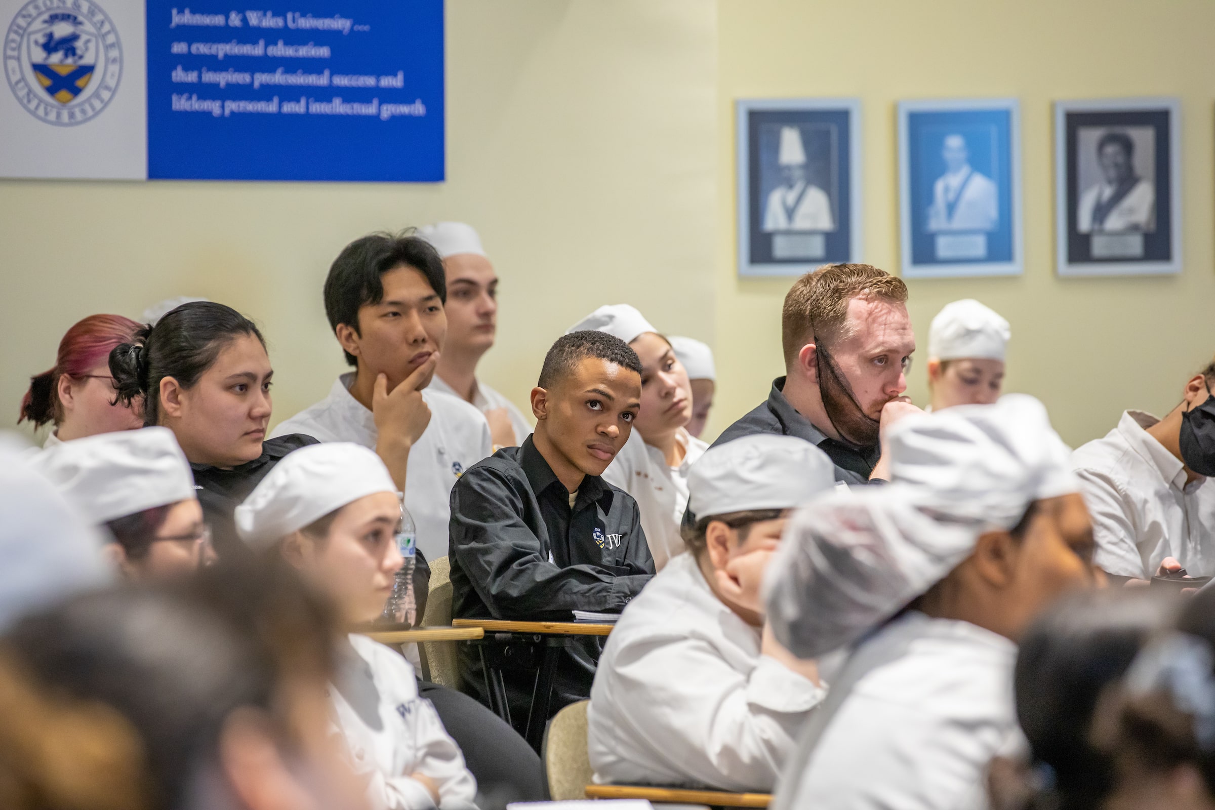 Group of culinary students in white jackets and watch caps listen to a chef giving a talk.