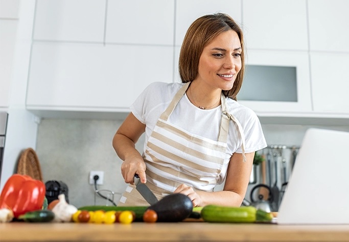 Female student chops vegetables on a cutting board while watching a culinary demonstration on her laptop.