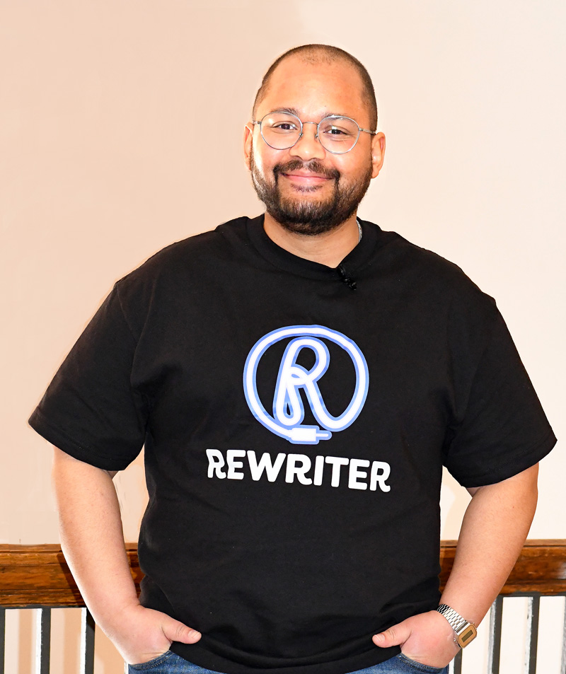 A smiling man wearing glasses and a black T-shirt with a stylized “R” logo and the word “REWRITER” stands with his hands in his pockets against a light-colored wall.