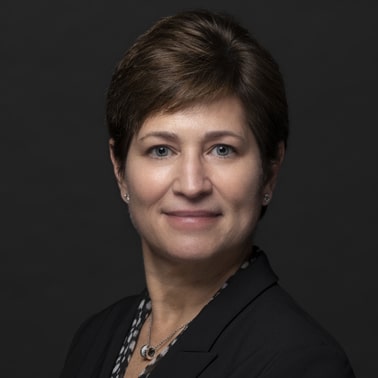 A professionally dressed individual with short brown hair and a subtle smile, set against a dark background.