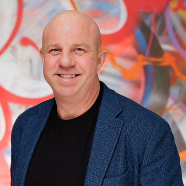A bald person with a warm smile wears a dark blue blazer over a black shirt, standing before a vibrant, abstract background.