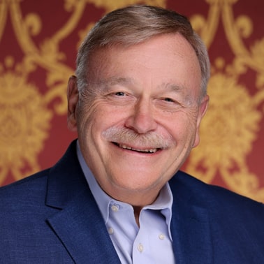 A smiling individual with a mustache, wearing a blue suit jacket and light blue shirt, stands against a red and gold ornate background.