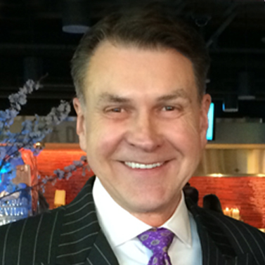 A person in a dark pinstripe suit and purple tie smiles warmly at the camera in an indoor setting.