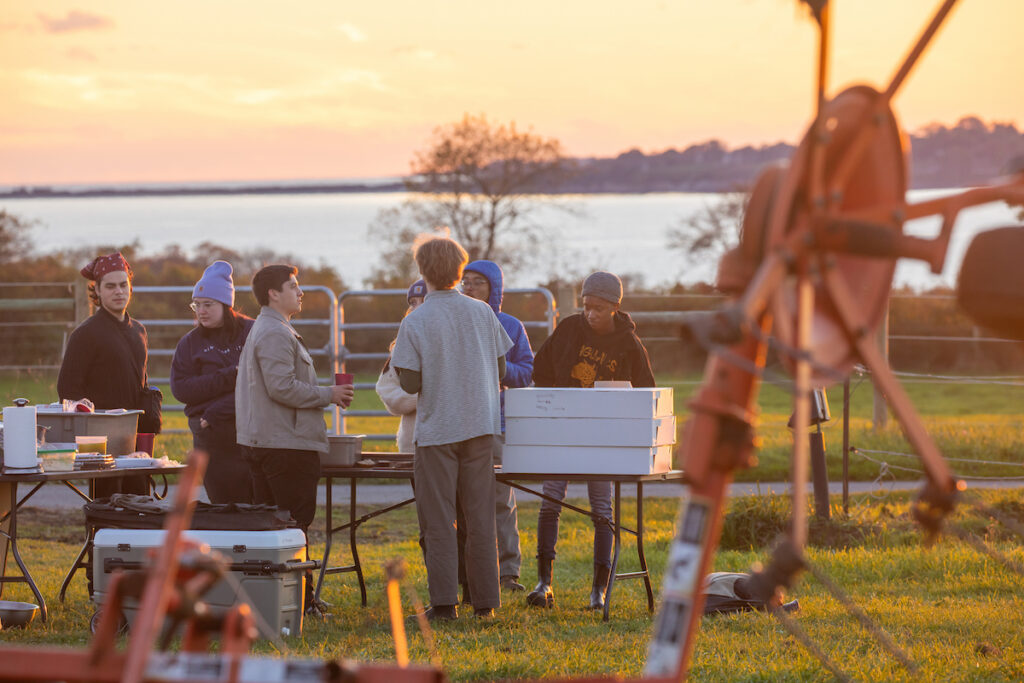 A group gathers outdoors at sunset near a body of water, surrounded by tables with supplies and equipment, with a large mechanical structure in the foreground.