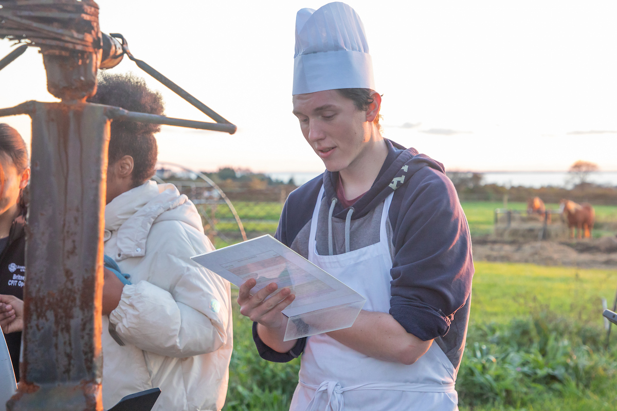 A person in a chef’s hat and apron holds a recipe sheet while standing outdoors near farm equipment, with others nearby and horses visible in the background.