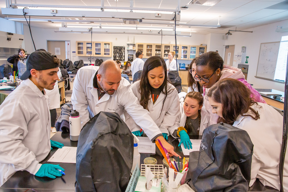 A group of students in lab coats and gloves gather around a table in a science lab, intently examining materials and discussing their findings with instructor.