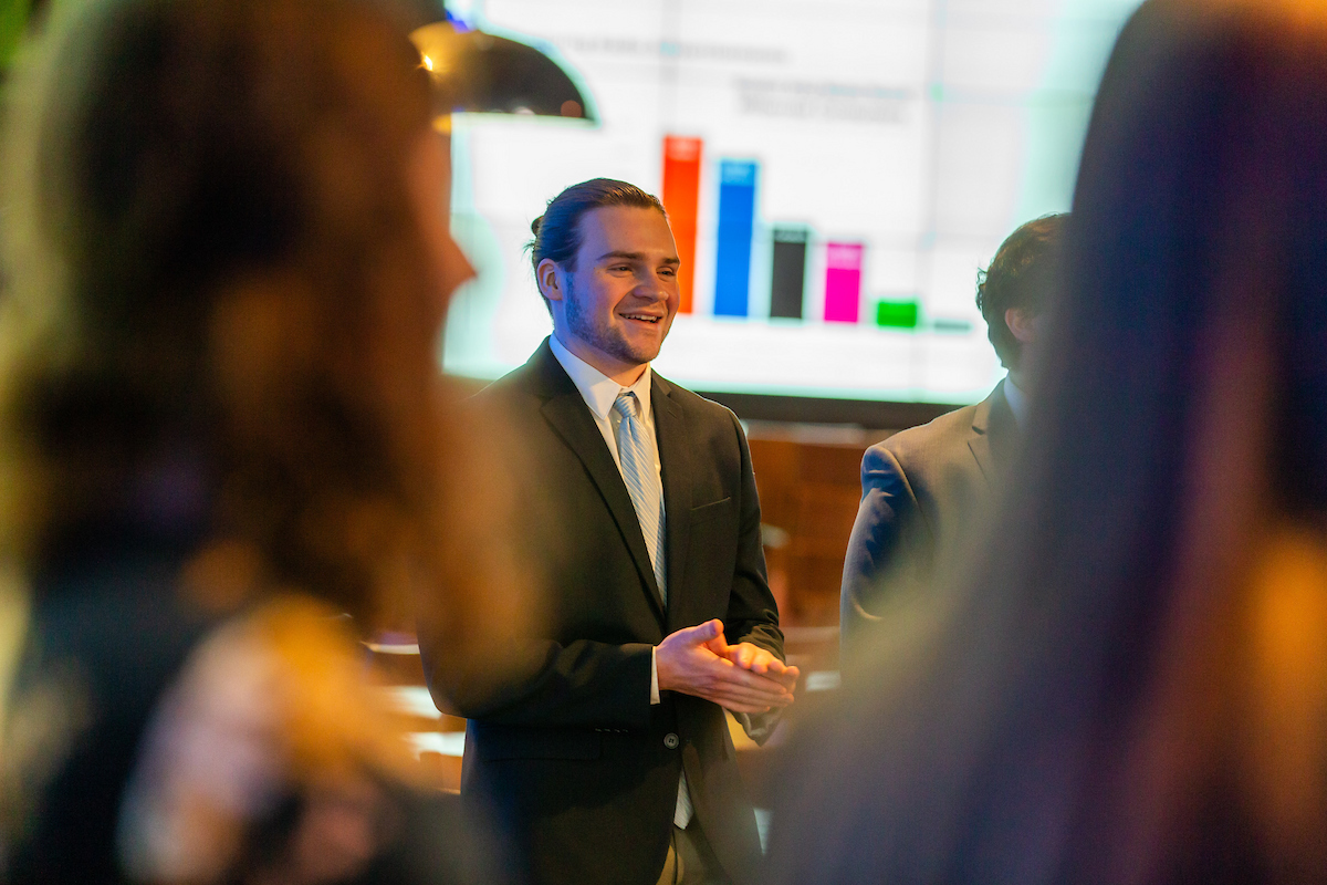 A person in a suit smiles while speaking to an audience during a presentation, with colorful bar charts visible on a screen behind them.