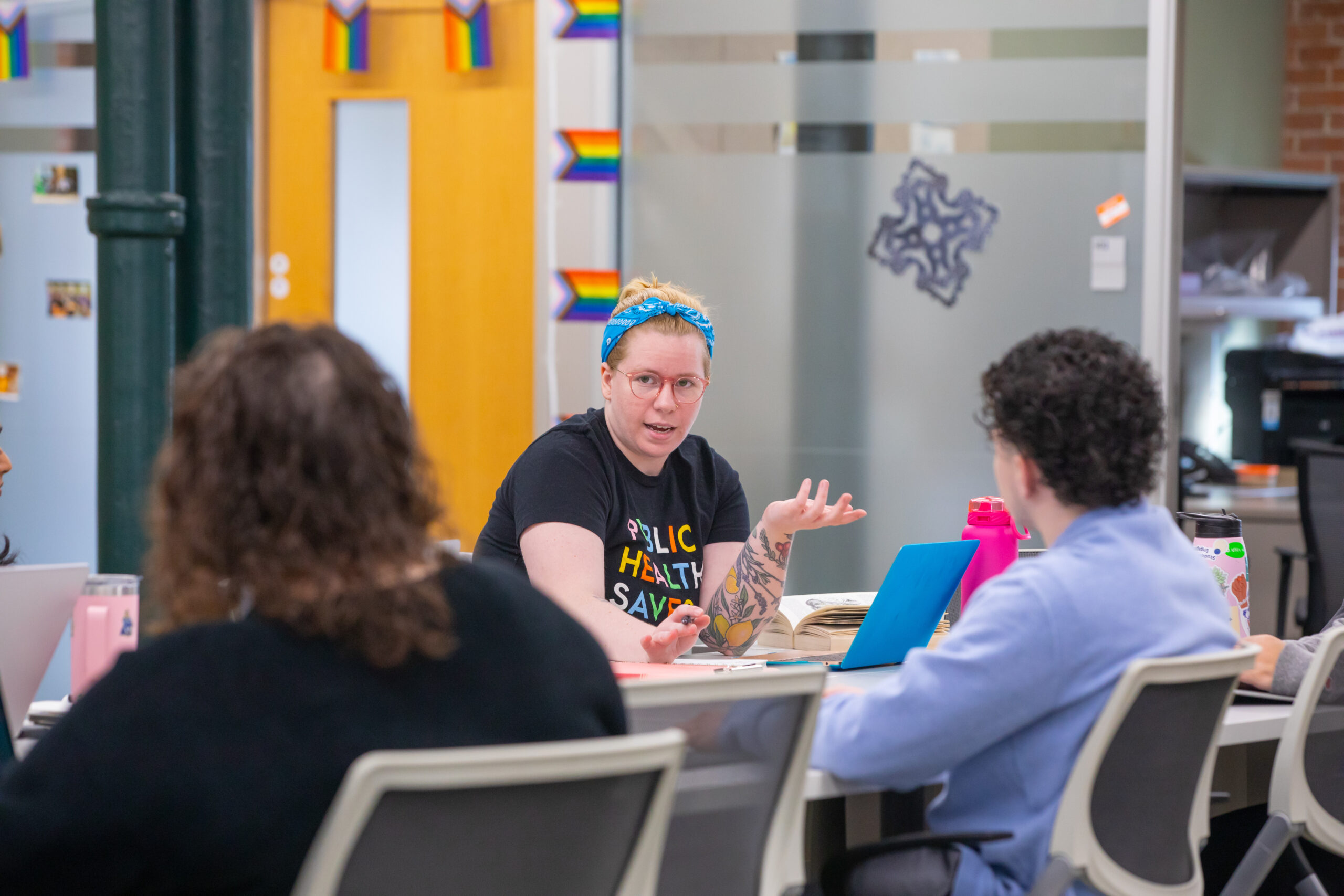 A person with a blue headband and tattoos gestures while speaking at a table during a meeting, surrounded by others in an office space decorated with rainbow flags.