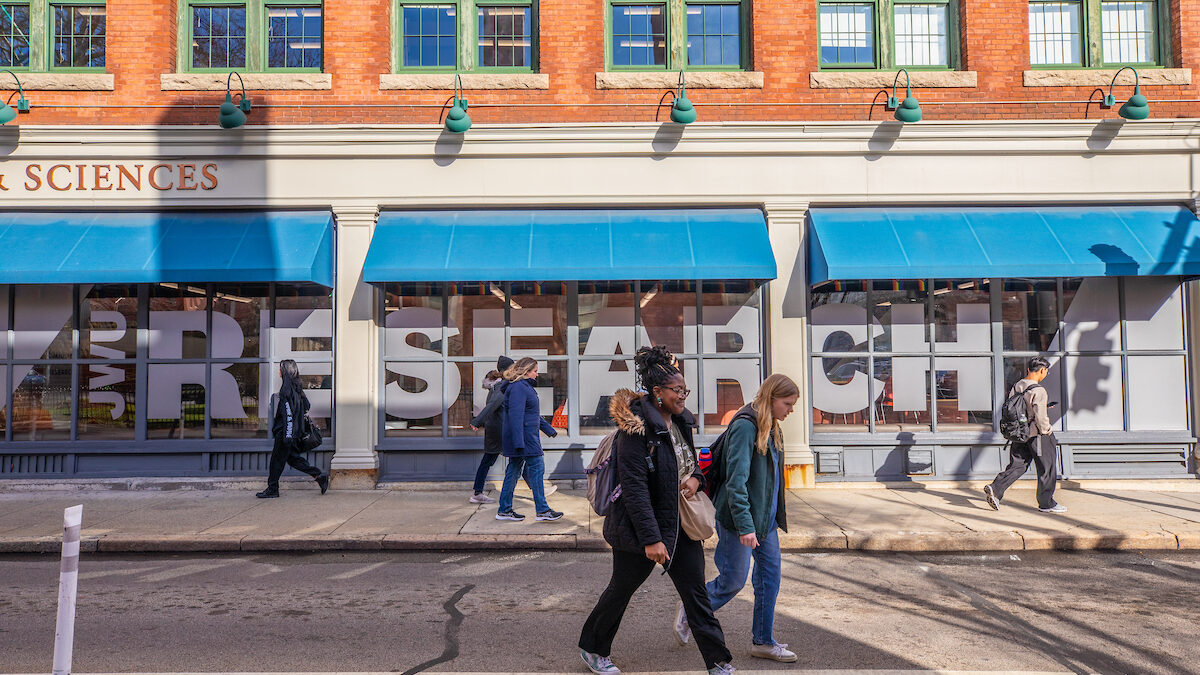 People cross a street in front of a brick building with blue awnings and large window displays featuring the word "RESEARCH."
