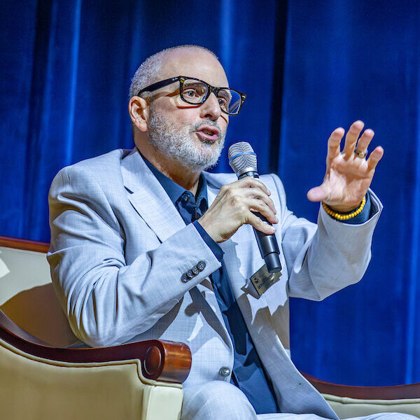 A person in a light gray suit and glasses speaks into a microphone while gesturing with their hand, seated in an armchair against a blue curtain backdrop.