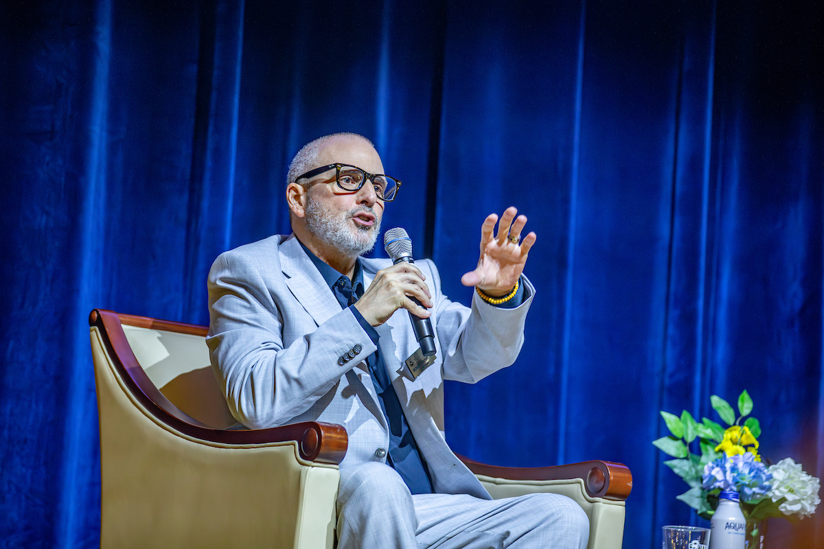 A person in a light gray suit and glasses speaks into a microphone while gesturing with their hand, seated in an armchair against a blue curtain backdrop.