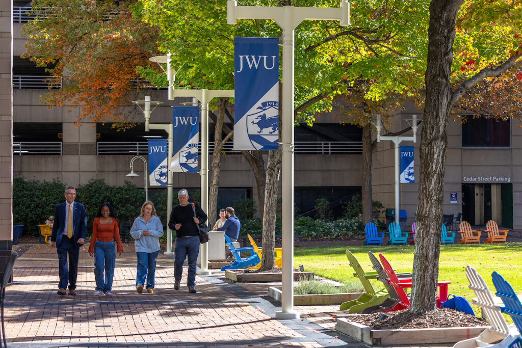 People walk along a brick pathway lined with trees and colorful chairs, passing by blue banners displaying "JWU" on poles, with a parking structure visible in the background.
