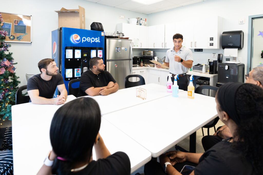 A person stands at the head of a table addressing a group seated around it in a break room with a Pepsi machine and kitchen appliances visible.