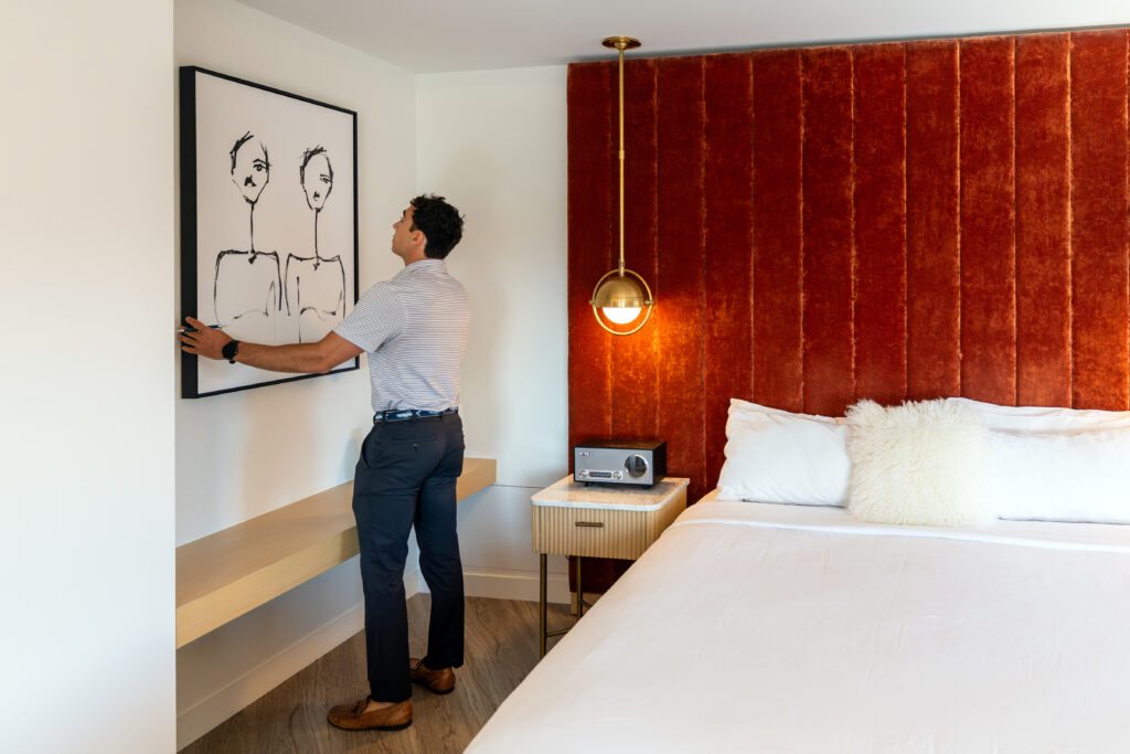 A person adjusts a framed artwork on a wall in a modern bedroom featuring a bold red velvet headboard and minimalist furnishings.