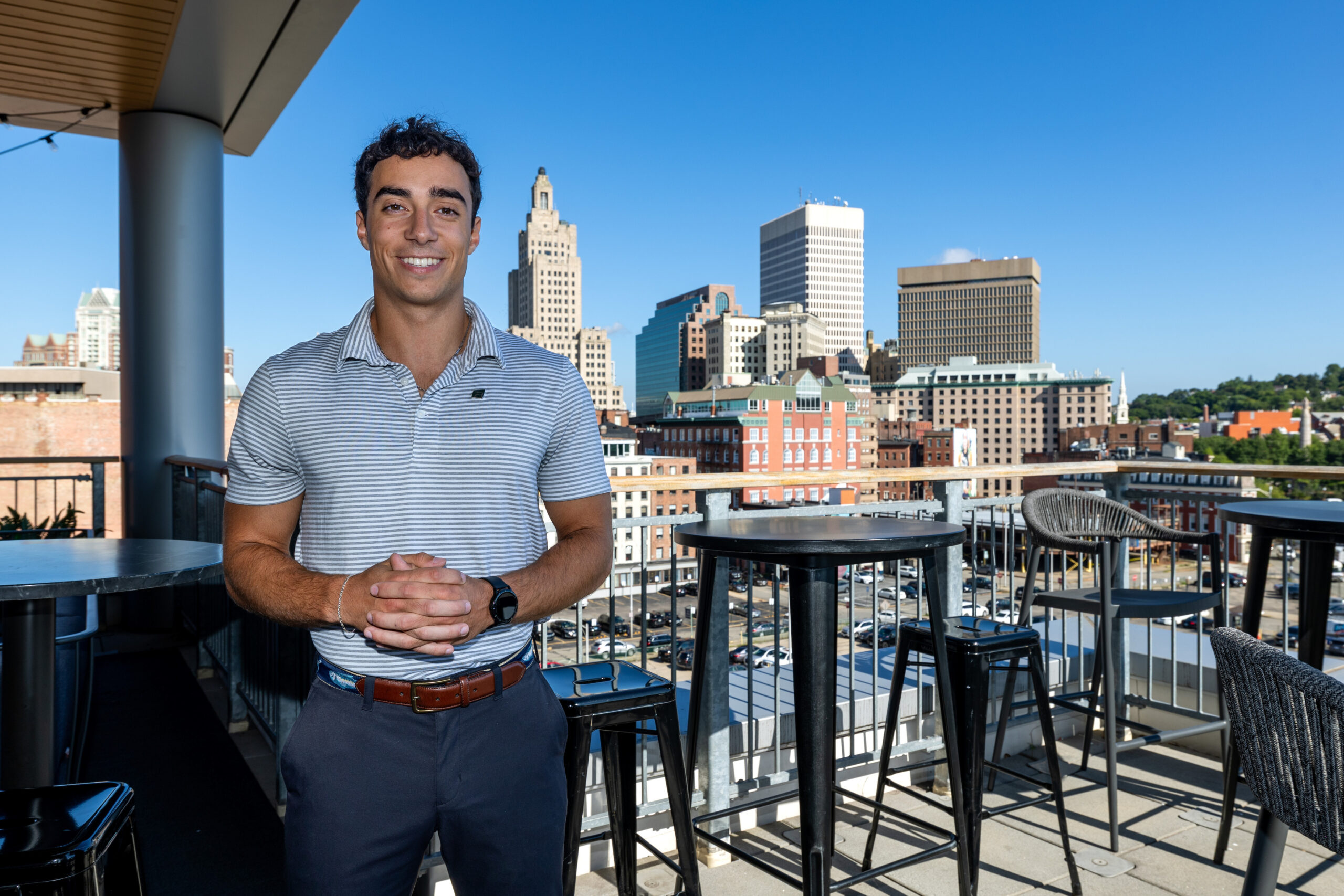 A person stands smiling on a rooftop patio with a city skyline in the background.
