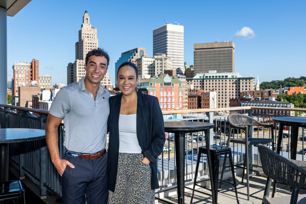 Two individuals smile for the camera on a rooftop patio with a city skyline in the background.