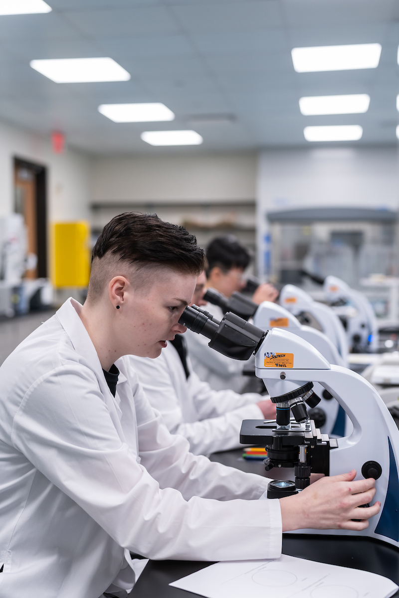 A person in a lab coat examines a specimen through a microscope in a well-lit laboratory.