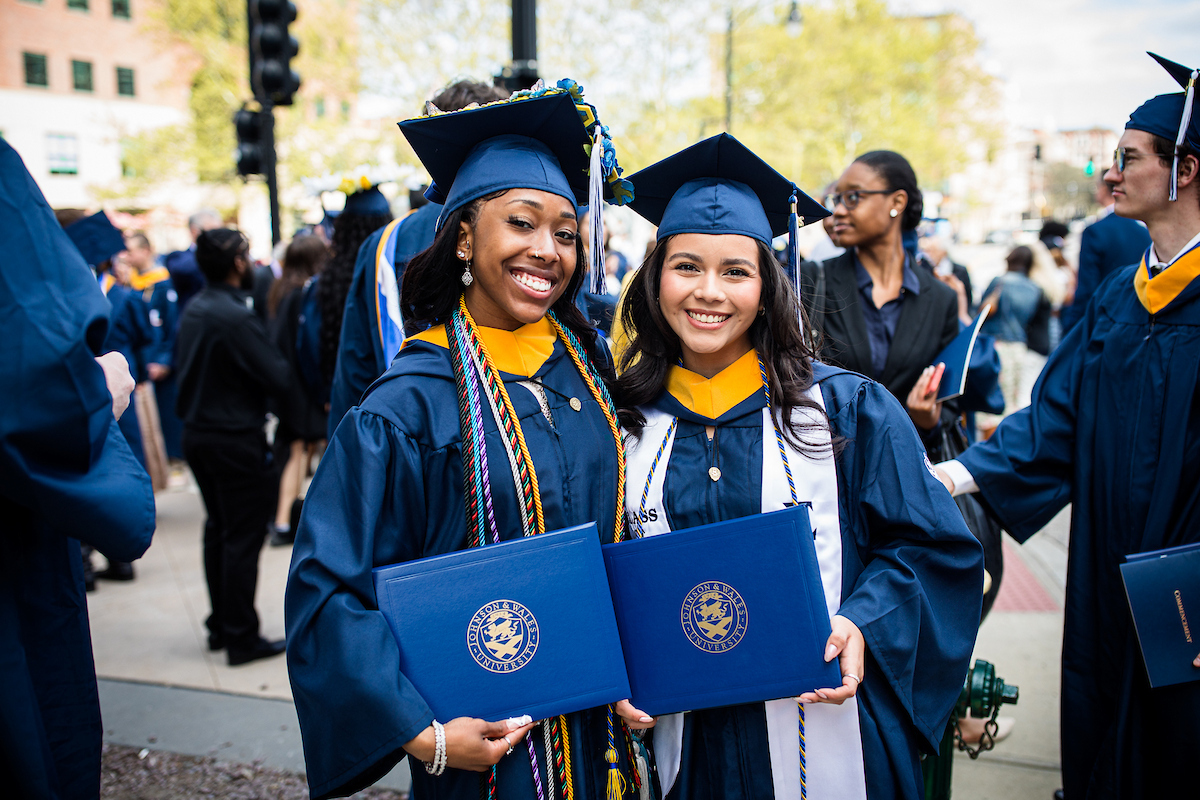 Two graduates in blue caps and gowns smile while holding diplomas from the University of Dayton during a commencement ceremony.