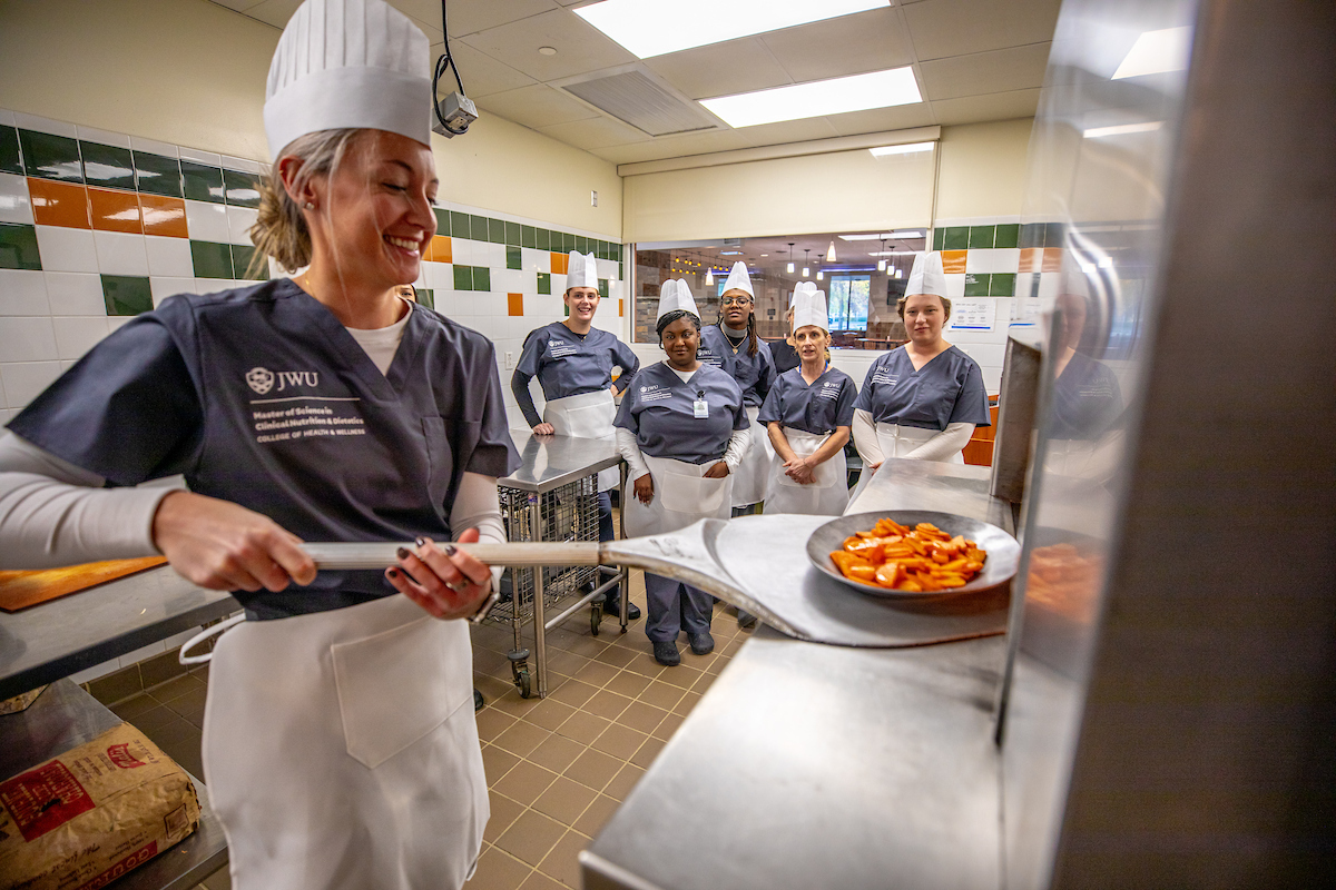 A group of culinary students in chef hats and uniforms stand in a kitchen, with one in the foreground smiling while handling a pan of cooked vegetables.