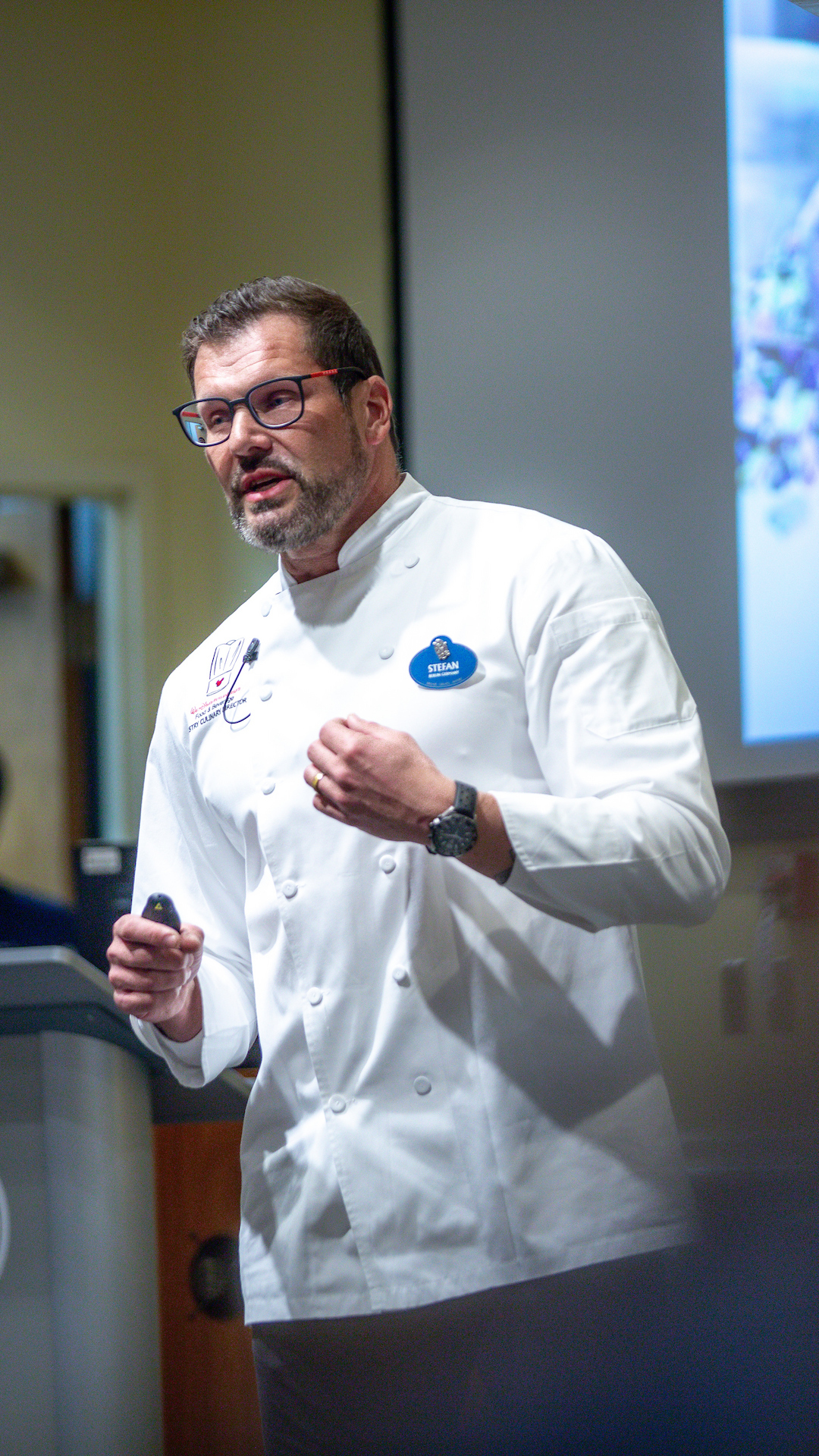 A chef in a white coat stands at a podium, speaking to an audience during a presentation at Johnson State University.