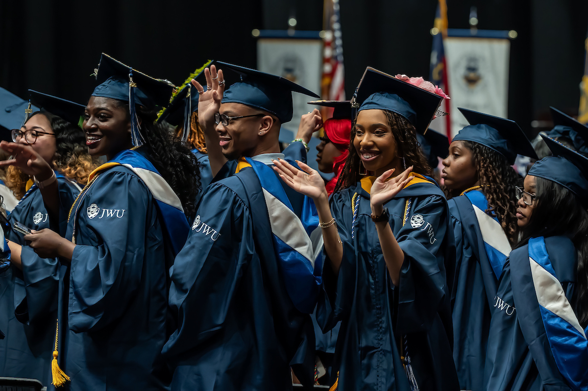 Graduates in blue and white gowns and caps wave and smile during a commencement ceremony.