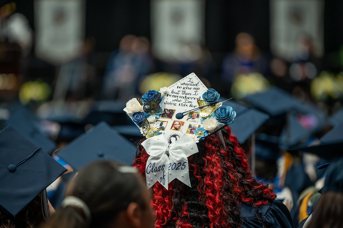 A graduate with vibrant red curls wears a decorated cap featuring a quote, photos, and a "Class of 2025" bow, standing among others in a graduation ceremony.