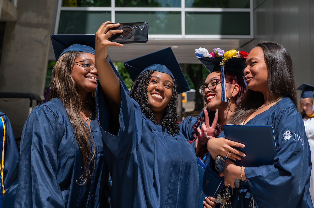 Graduates in blue caps and gowns take a selfie together outdoors, smiling and posing with graduation caps and diplomas.