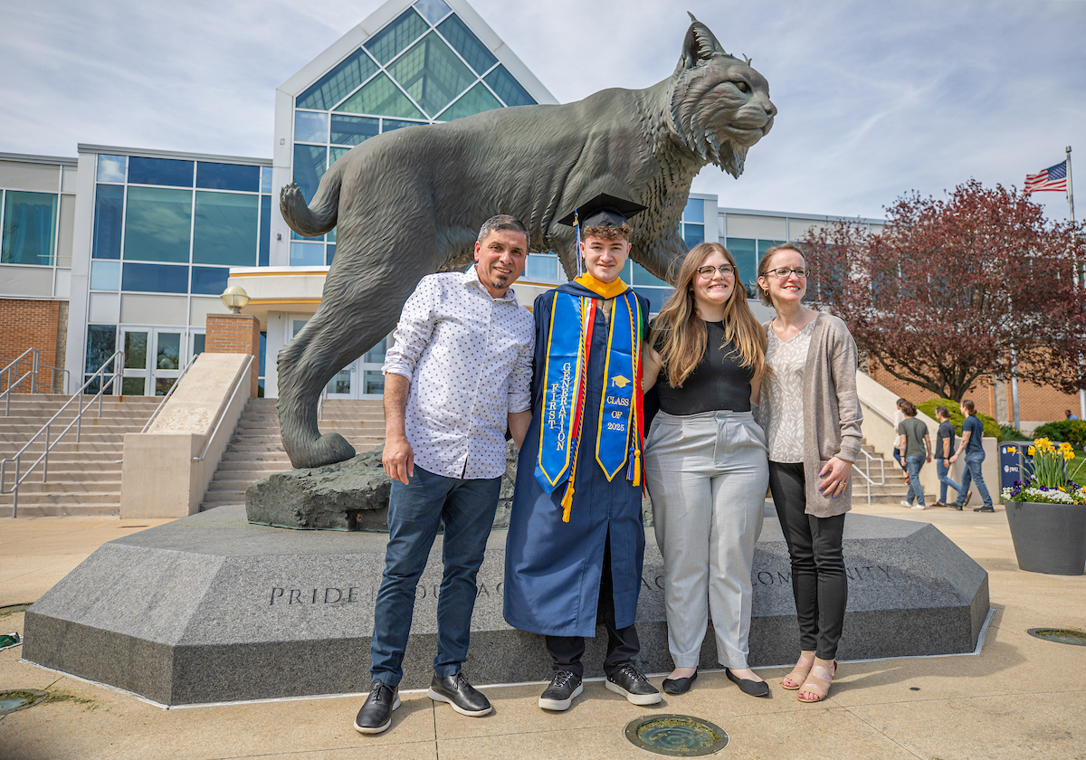 A graduate in a cap and gown stands with three adults in front of a large statue of a lynx on a campus.