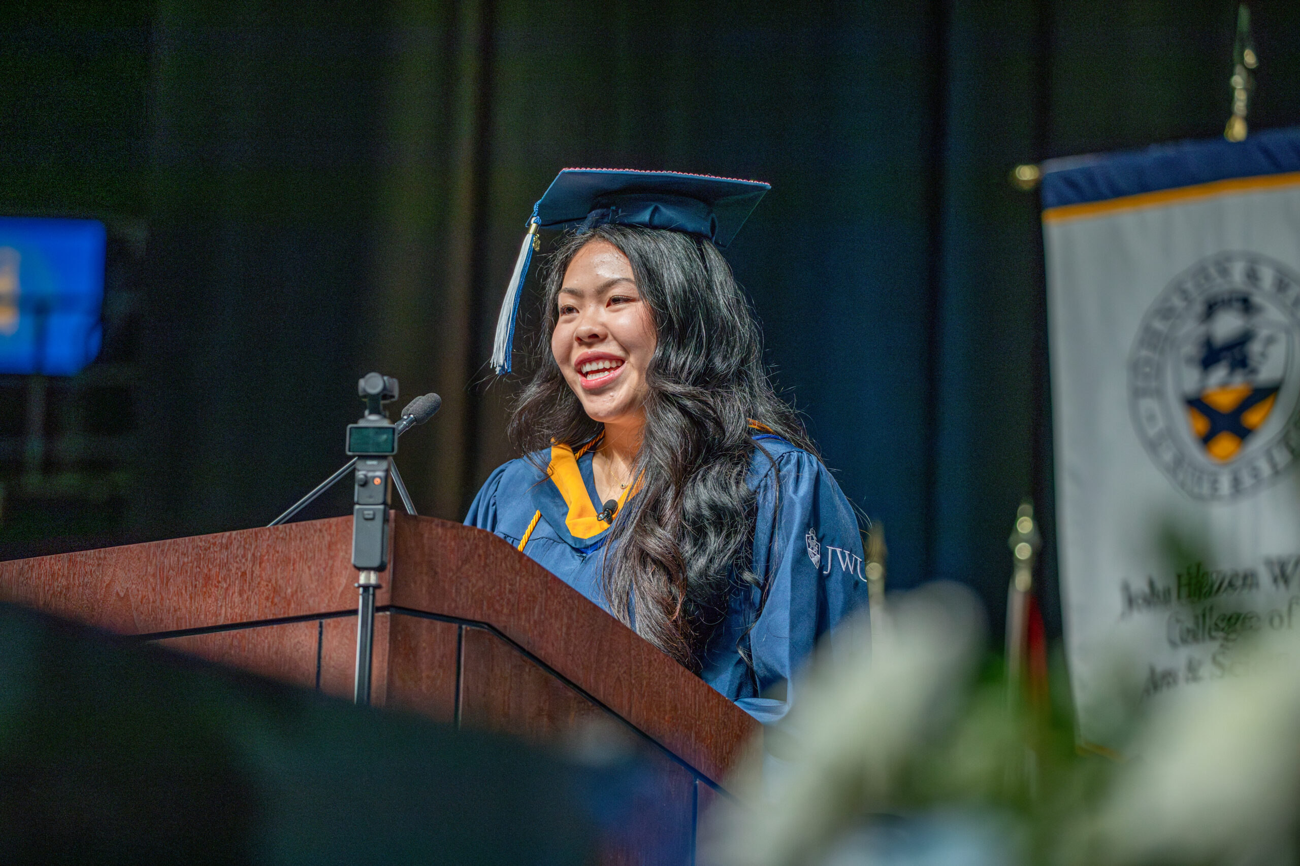 A graduate in a blue cap and gown smiles while speaking at a podium during a ceremony.