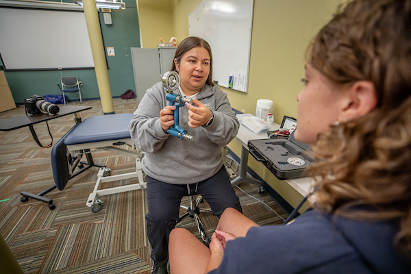 A person in a gray sweatshirt demonstrates the use of a blue hand grip tool to another individual seated on an examination table in a clinical setting.
