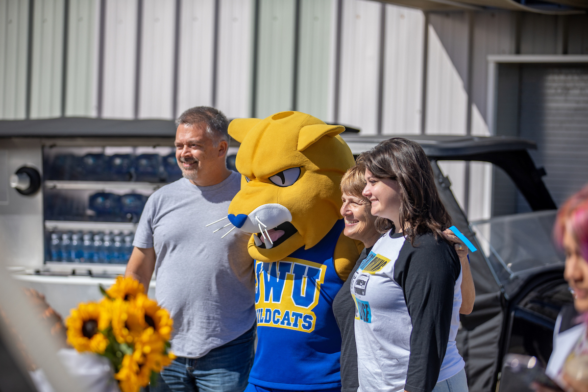 A yellow mascot wearing a blue "OWU Wildcats" jersey stands with two people outdoors near a vehicle and a cooler filled with water bottles.