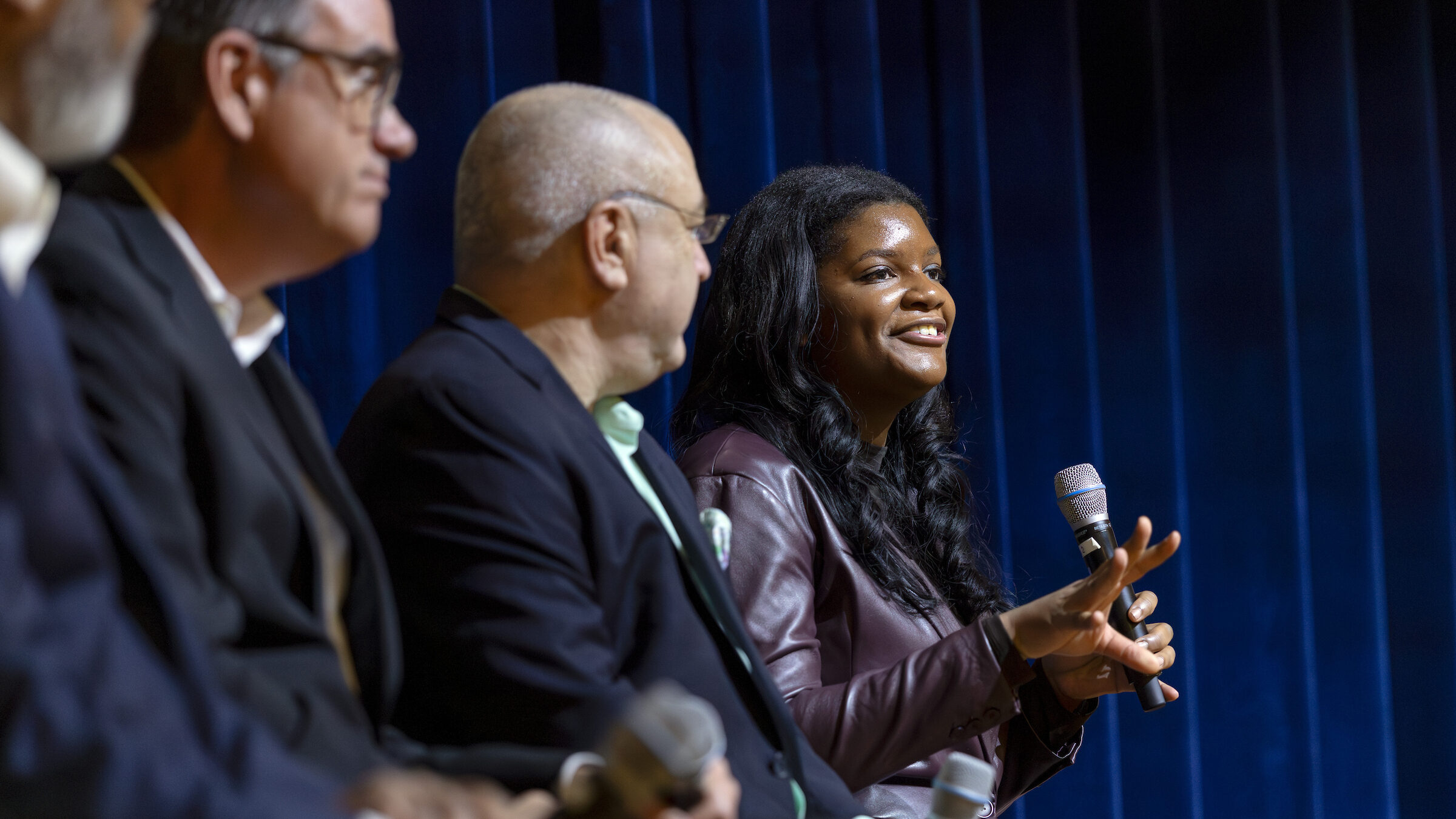 A panel of four individuals seated on stage, with one person speaking into a microphone while the others listen attentively against a blue curtain backdrop.