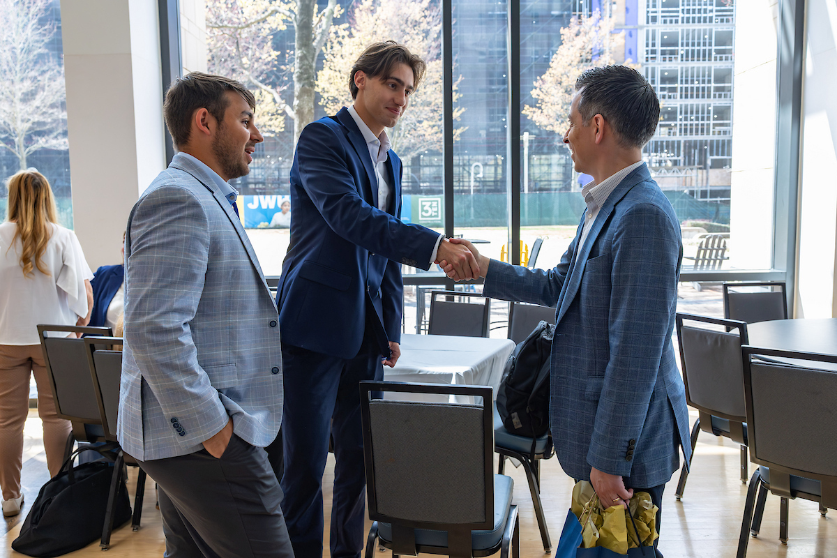 Three individuals in business attire shake hands in a modern, well-lit indoor space with large windows and tables.
