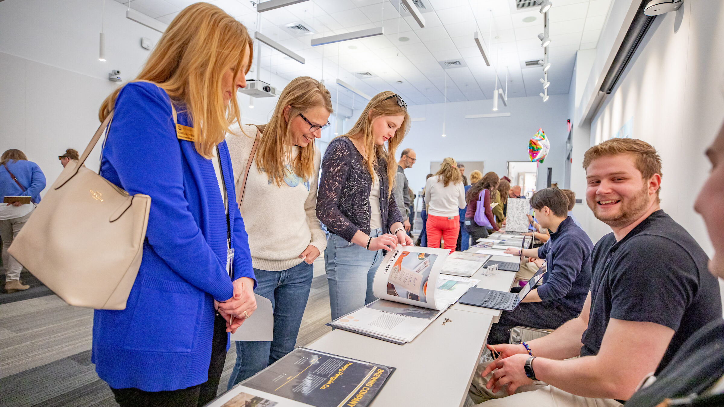 A group of people browse materials at a long table in a brightly lit indoor space, with some seated and others standing.