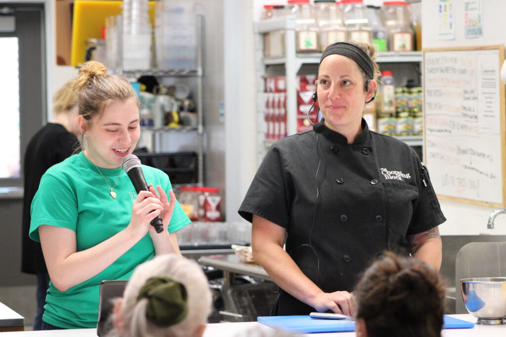 Ava Zajczenko, speaking into a microphone, introduces that month's special guest while leading a cooking class at the Common Pantry
