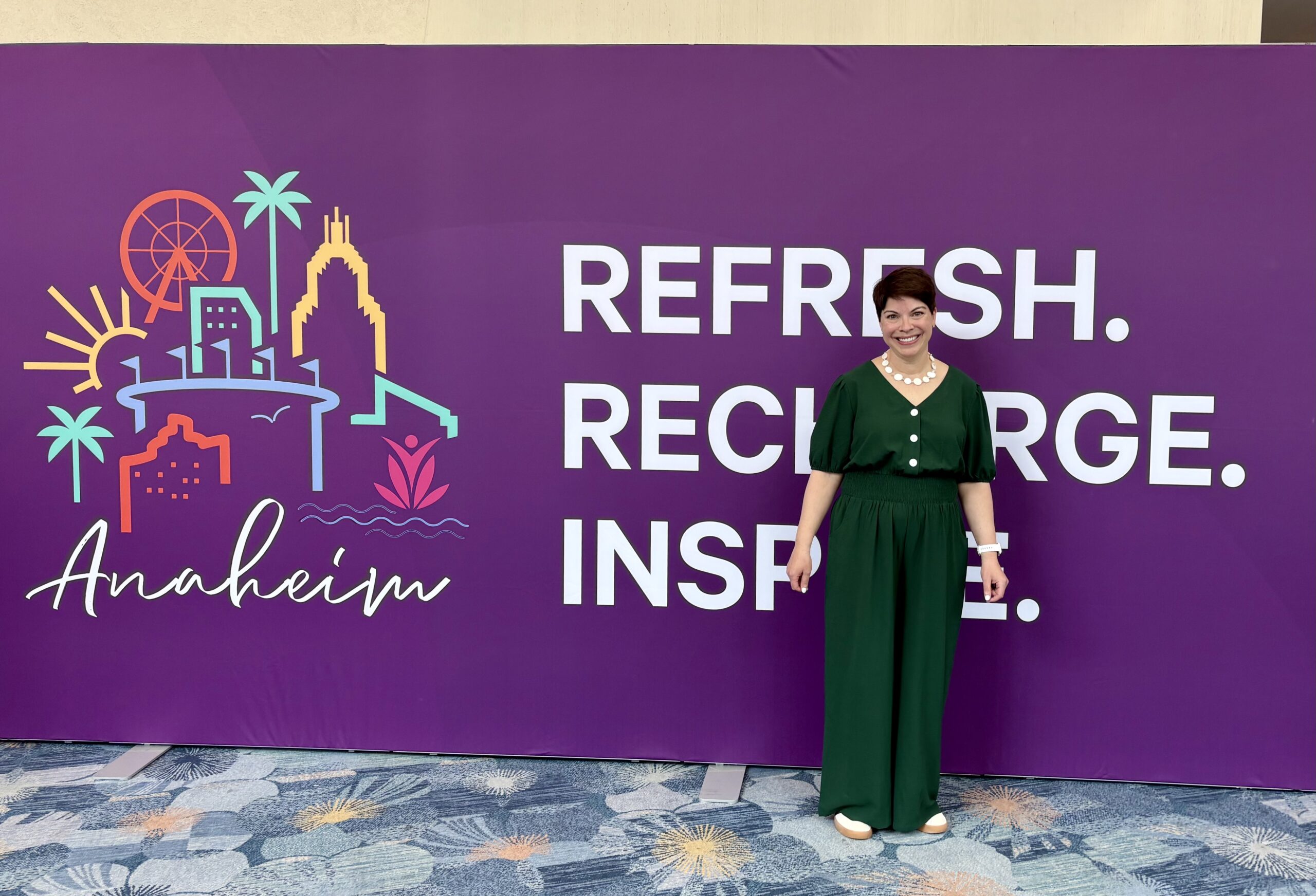 a nicely dressed woman stands smiling against a mural for the Inspire 2026 conference