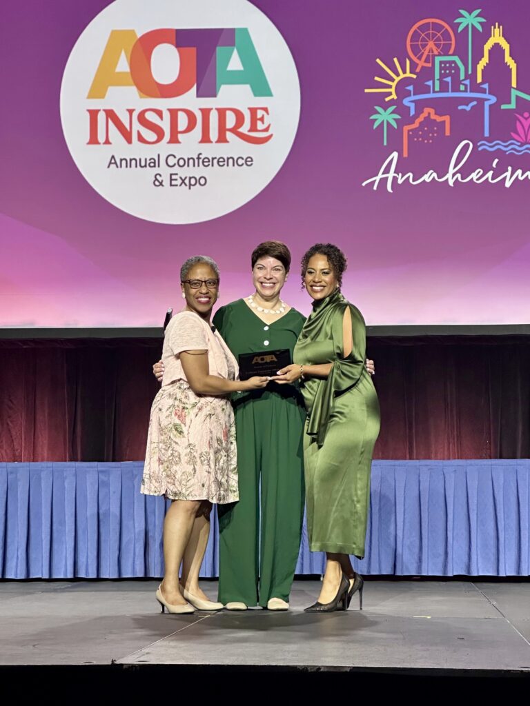 three women pose together on a stage, surrounding an award, against a backdrop for the AOTA Inspire conference