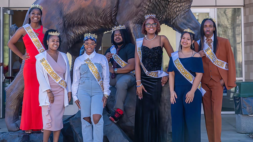 Group of seven college homecoming court members wearing crowns and “Homecoming Court” sashes, posing together in front of a large bronze wildcat statue.