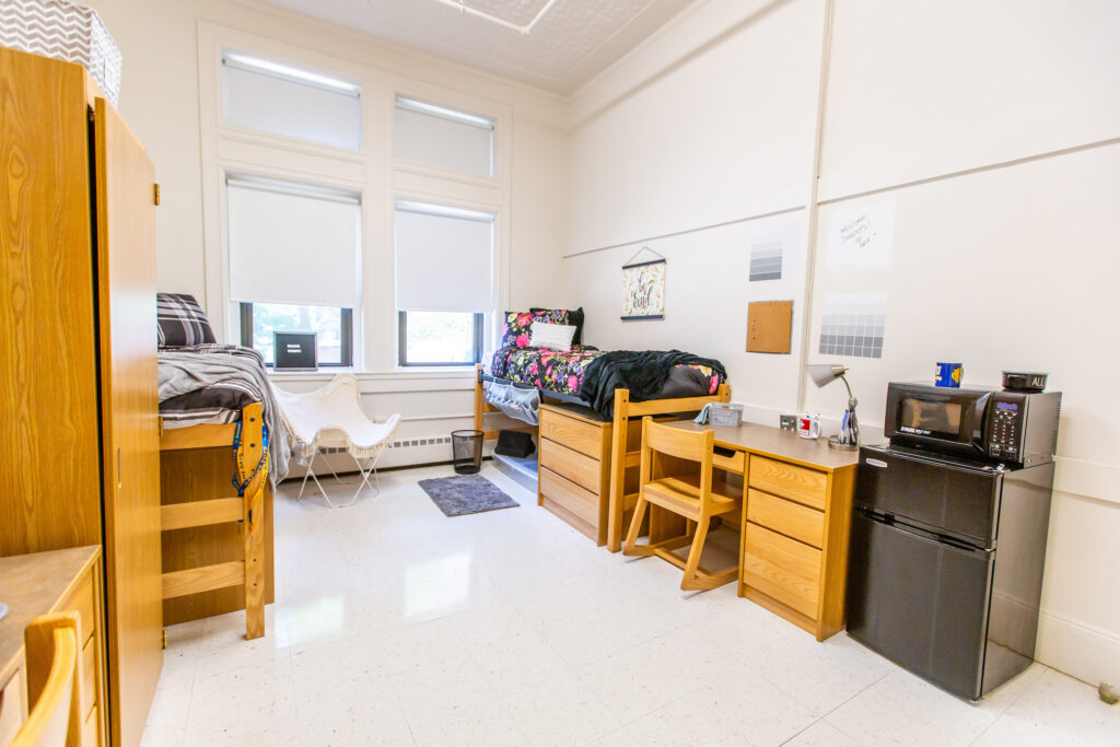 A clean, well-lit dorm room with two wooden bunk beds, a desk with a chair, a small refrigerator, a microwave, and large windows with blinds.
