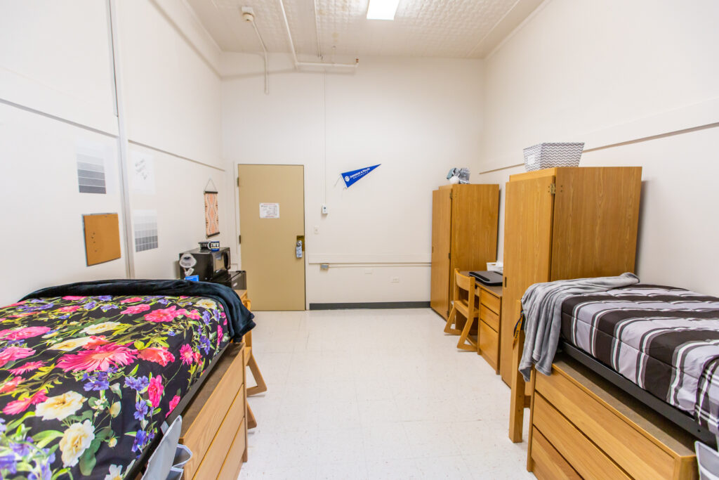 A clean, minimalist dorm room with two wooden beds, matching desks and dressers, and a beige door at the far end.