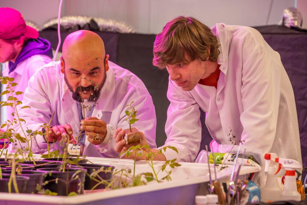 Two individuals in lab coats examine small plants in pots under purple lighting.