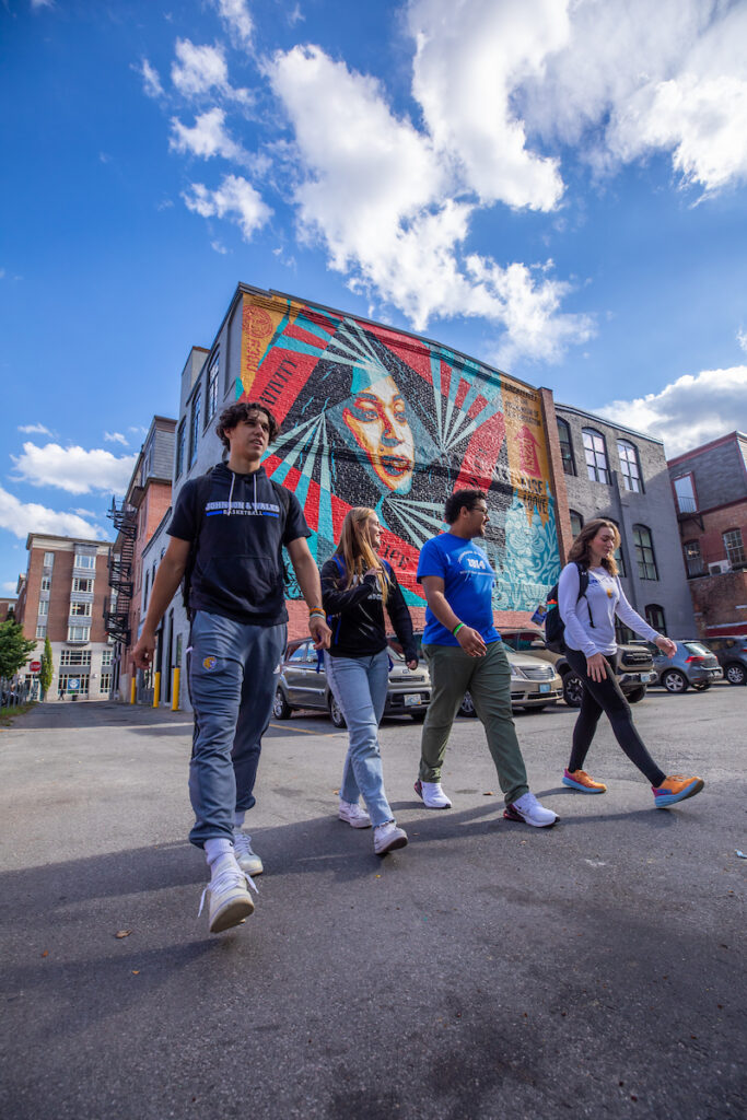 Four individuals walk across a street under a bright blue sky, passing a large colorful mural on a brick building.
