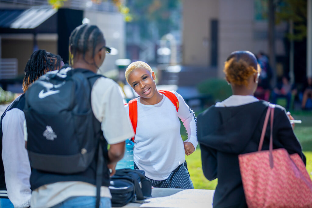 A group of young people gather outdoors, with one person smiling at the camera while others converse nearby.