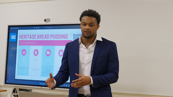 A person in a navy blazer stands before a presentation screen displaying "Heritage Bread Pudding" and business-related icons.