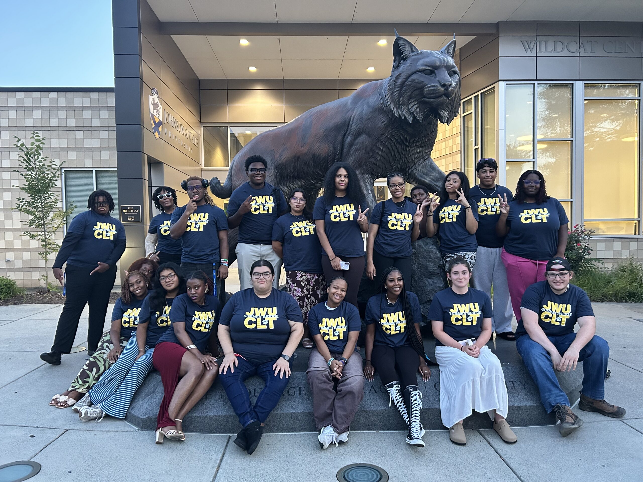 A large group poses outdoors in front of a dark bronze wildcat statue at the entrance to the Wildcat Center.