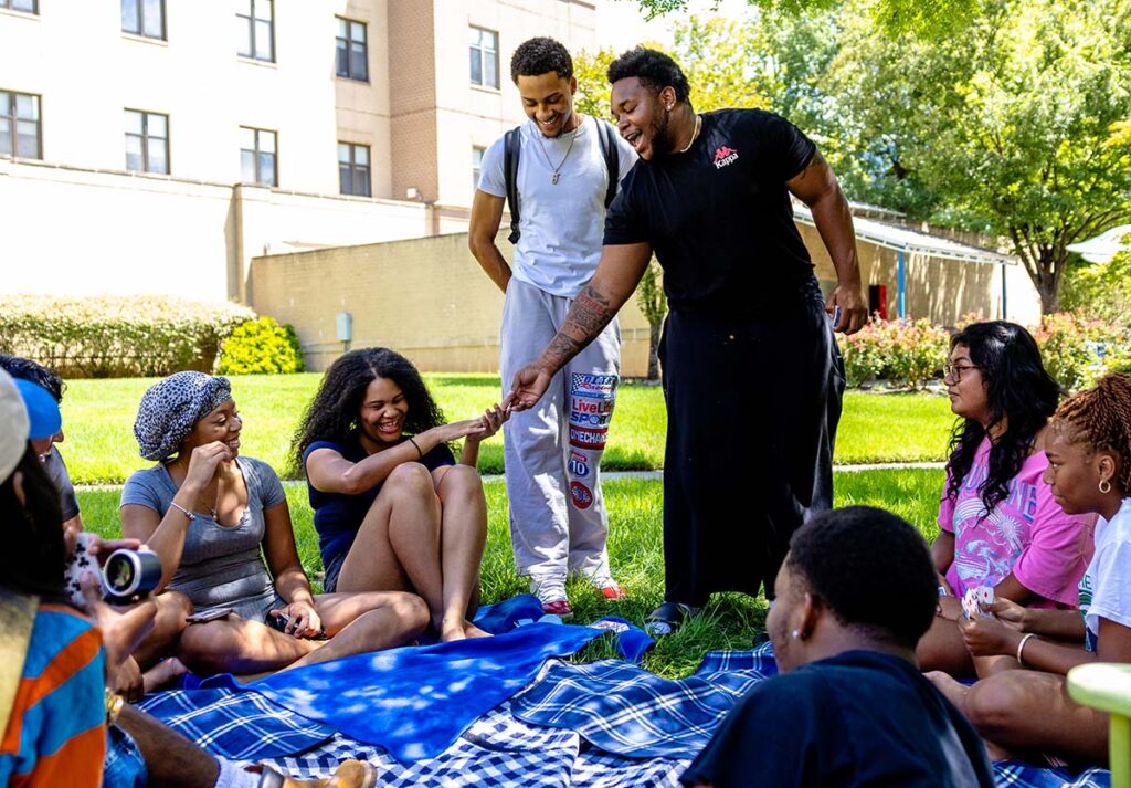 A group of people gathers outdoors on a sunny lawn, seated on blue and plaid picnic blankets while two people stand in the center. The standing person on the left wears a light gray shirt and sweatpants with colorful patches; the person on the right wears a black shirt and black pants and gestures toward a seated woman.