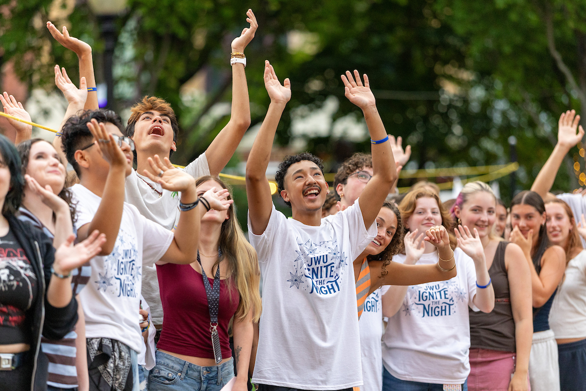 A group of young people wearing "JWU IGNITE THE NIGHT" shirts raise their hands in excitement during an outdoor event.