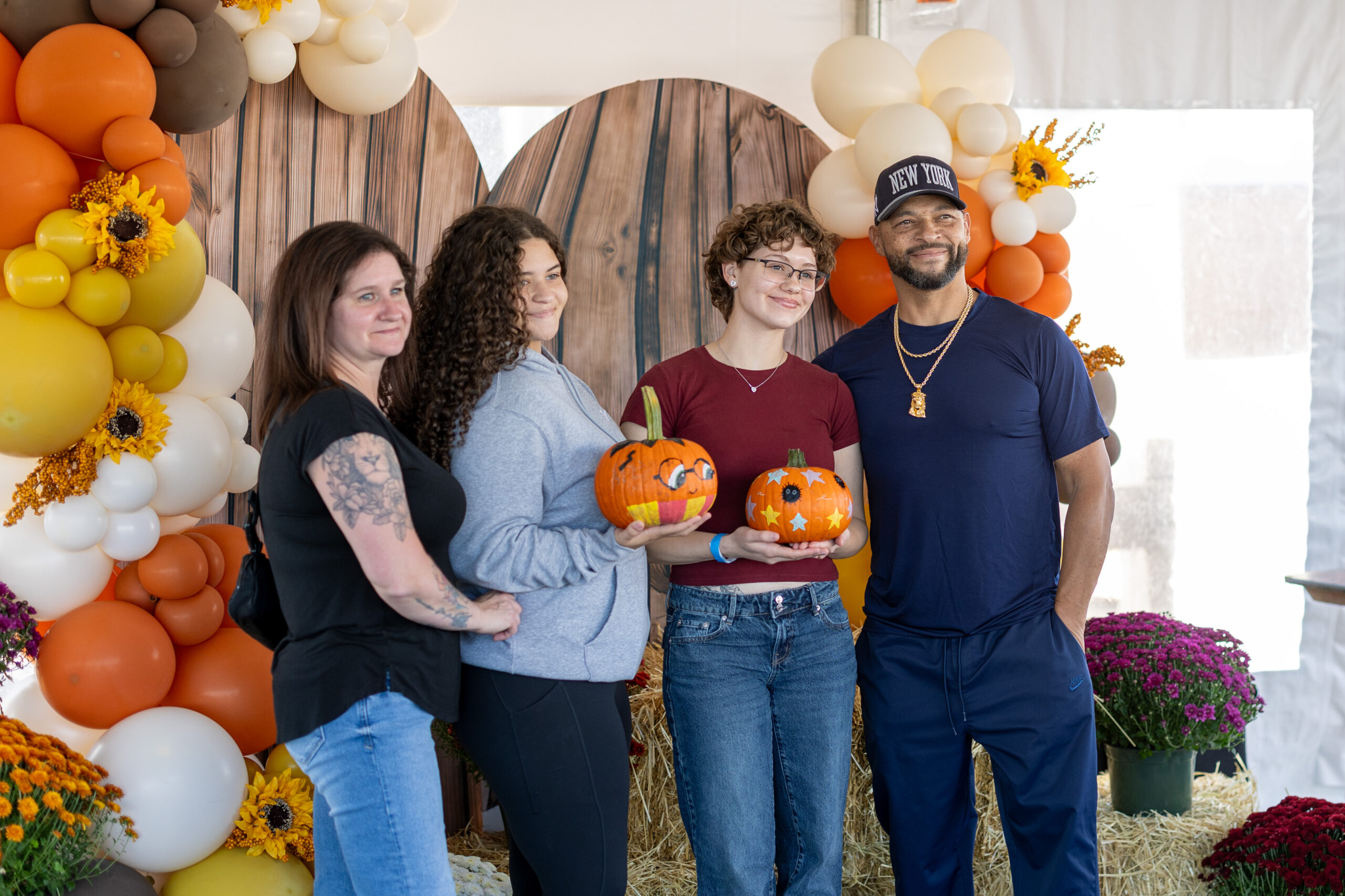 Four individuals stand together at an autumn-themed event, holding decorated pumpkins, surrounded by colorful balloon arches and seasonal flowers.
